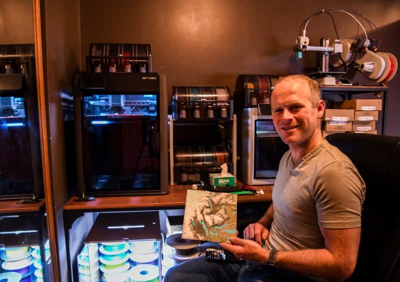 David in his workshop holding a topographic print, surrounded by 3D printers